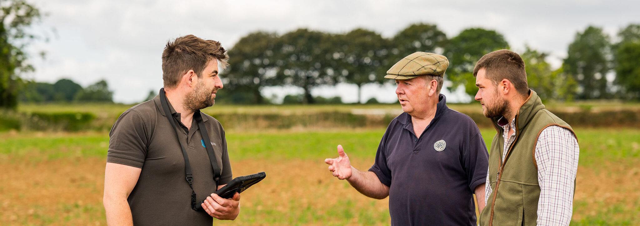 3 persons in a field talking with row of trees in background