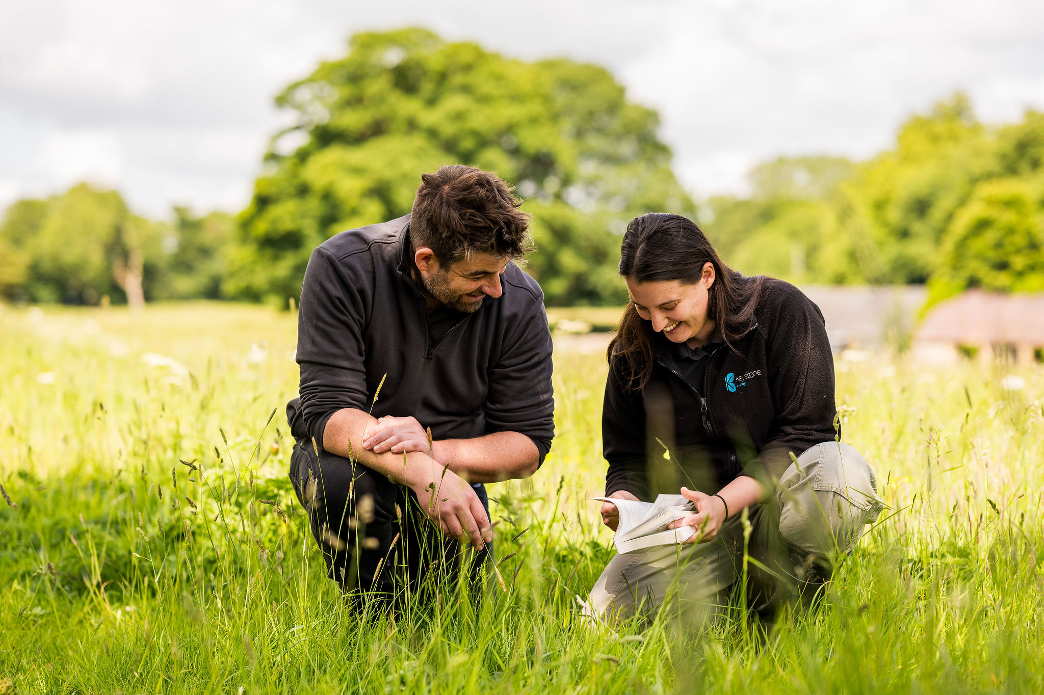 A man and a woman crouched down in a field looking at a book and inspecting grasses